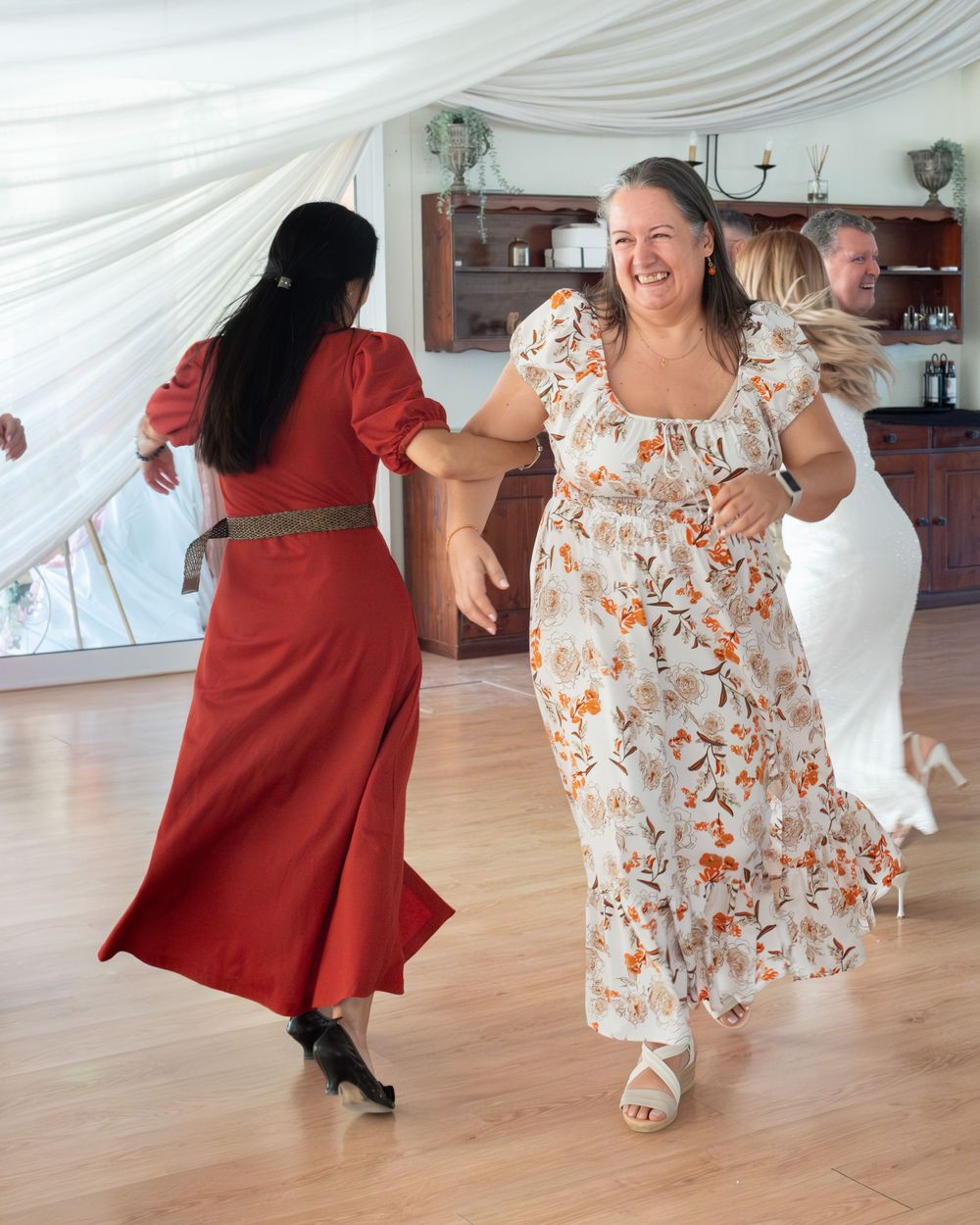 Two women in floral and red dresses dancing joyfully at an indoor celebration on a wooden floor.