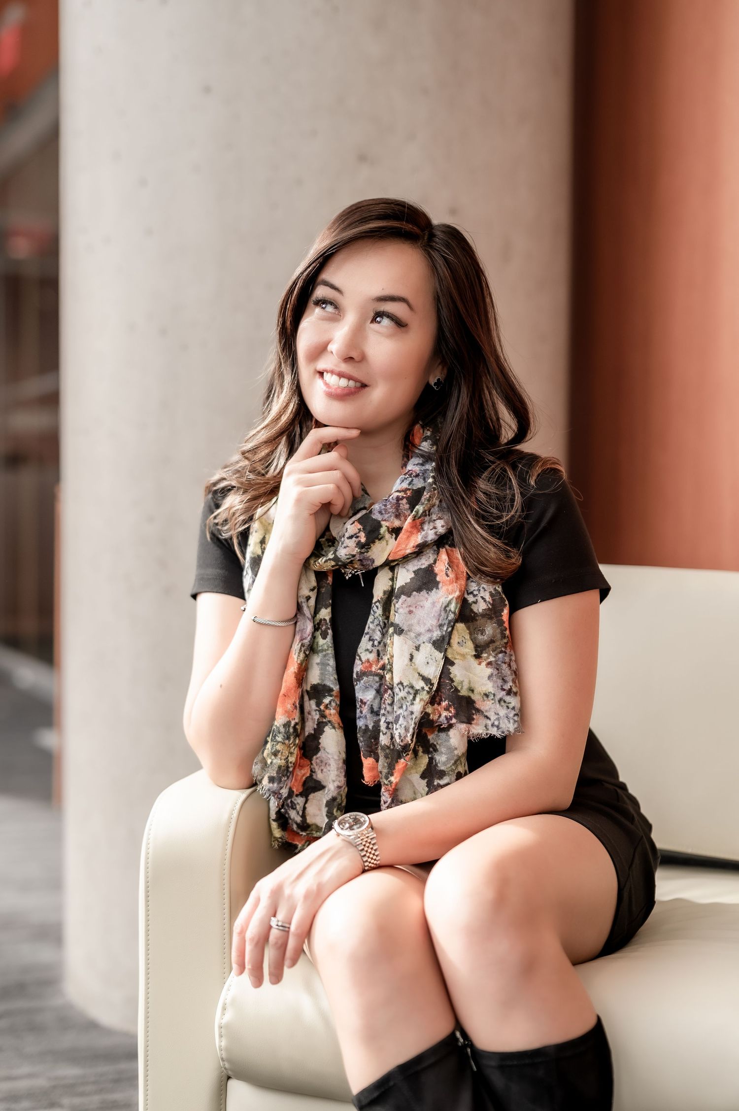 Smiling young woman in black dress and floral scarf sits cross-legged in cream chair, posing thoughtfully indoors.