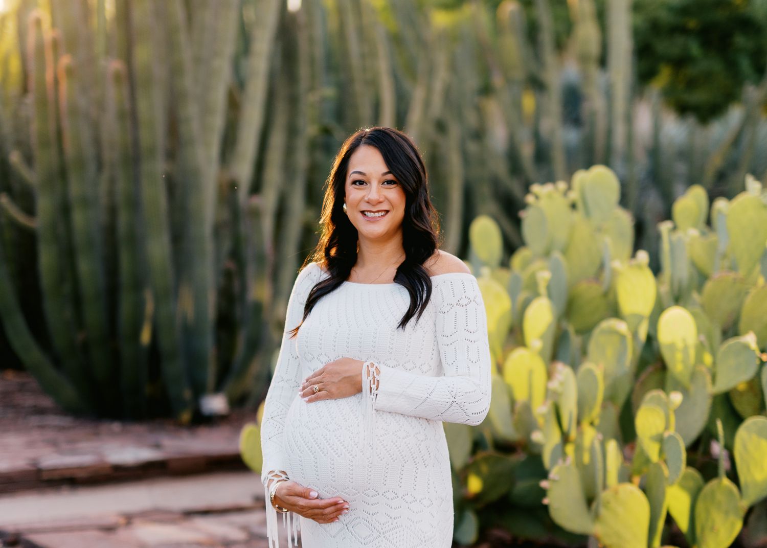 Pregnant woman in white lace dress smiling outdoors among tall cacti and prickly pear plants.