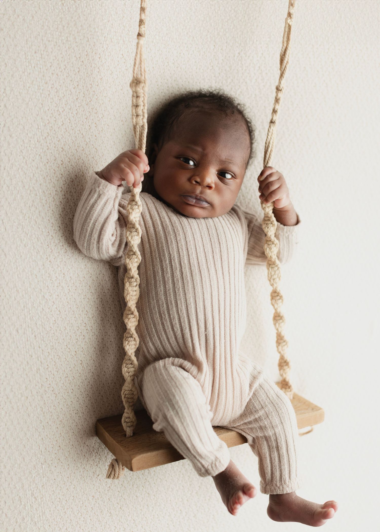 Adorable baby in a cream ribbed romper sitting on a wooden macrame swing against a white background. Elizabeth Lang Photography Surrey