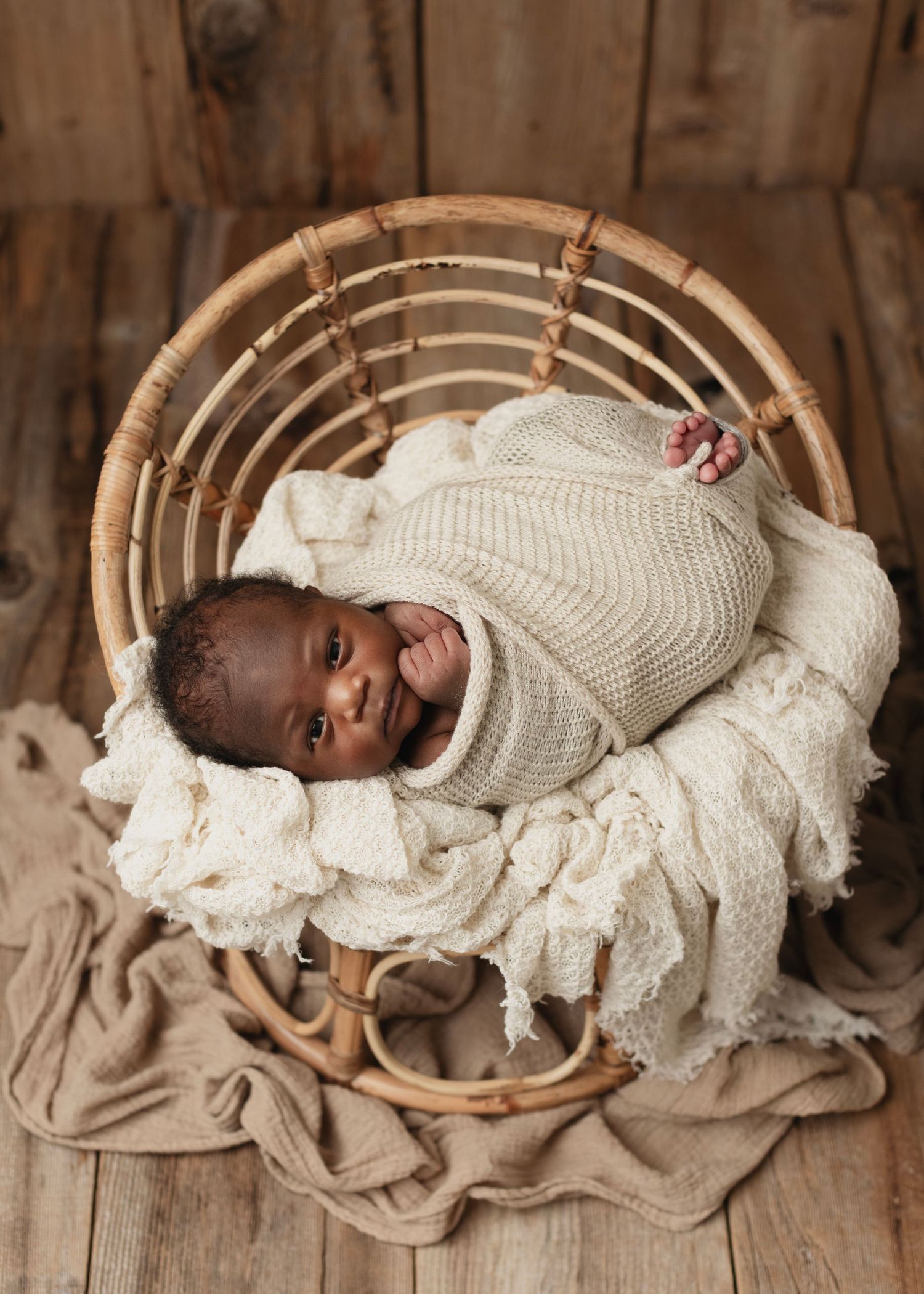 Smiling newborn baby wrapped in cream knit blanket resting in a wicker basket on wooden floor. Elizabeth Lang Photography Surrey