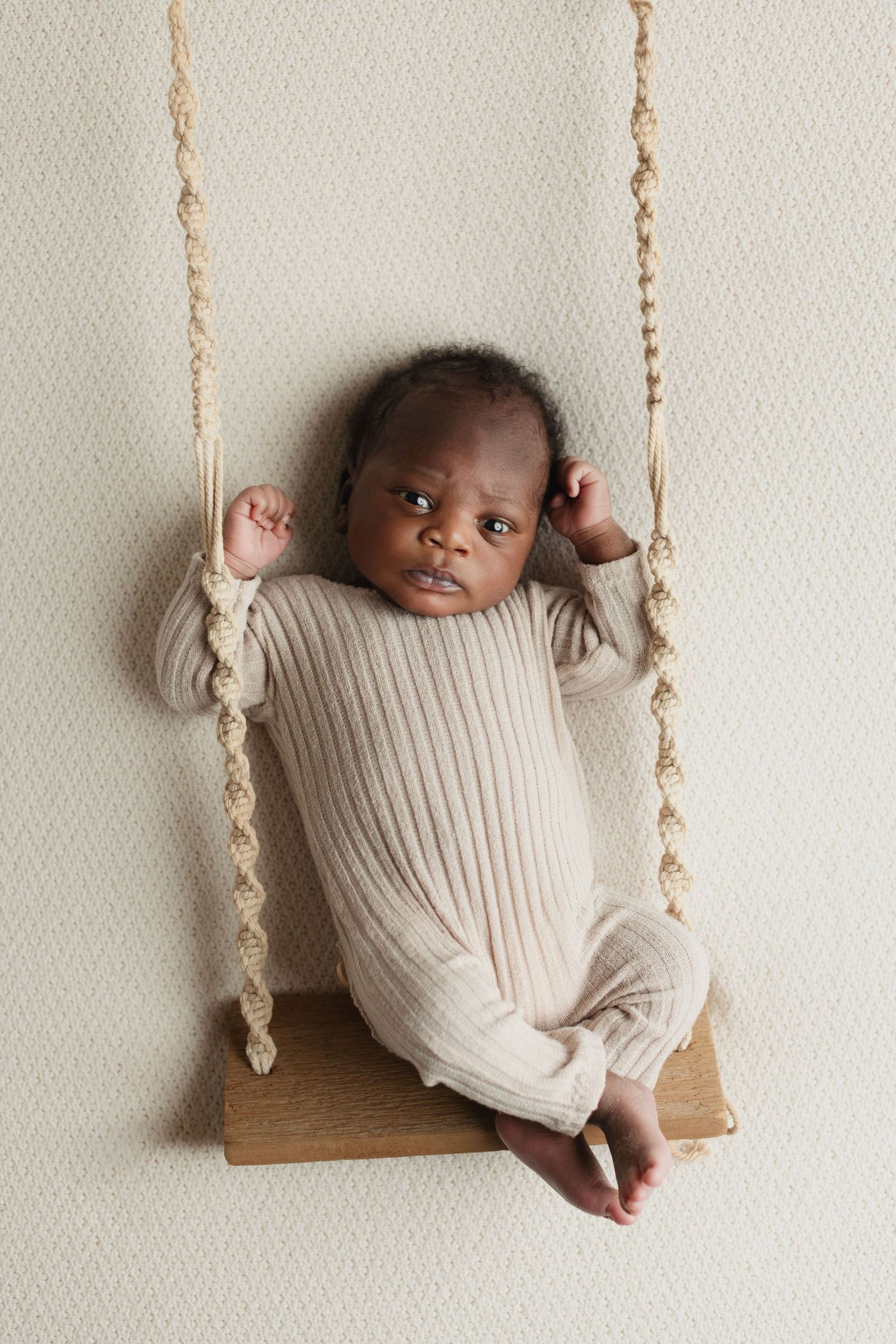 Newborn baby in beige ribbed onesie resting on a wooden swing with rope, against a neutral background. Elizabeth Lang Photography Surrey