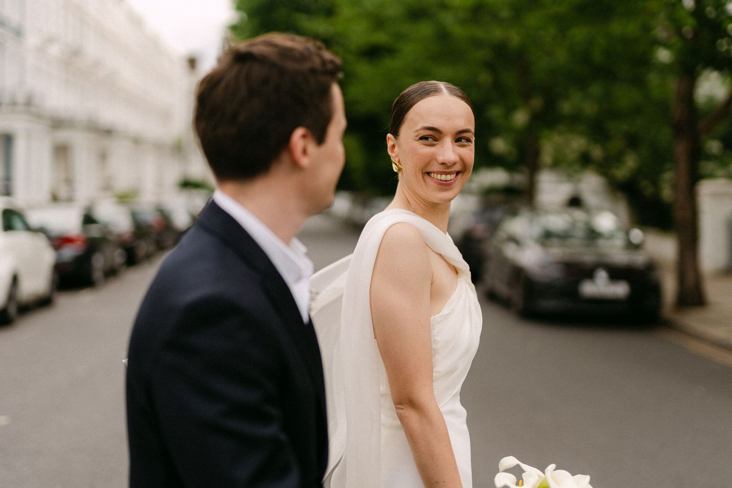 Bride smiling over shoulder at groom on a tree-lined street, wearing a white gown and holding a bouquet.