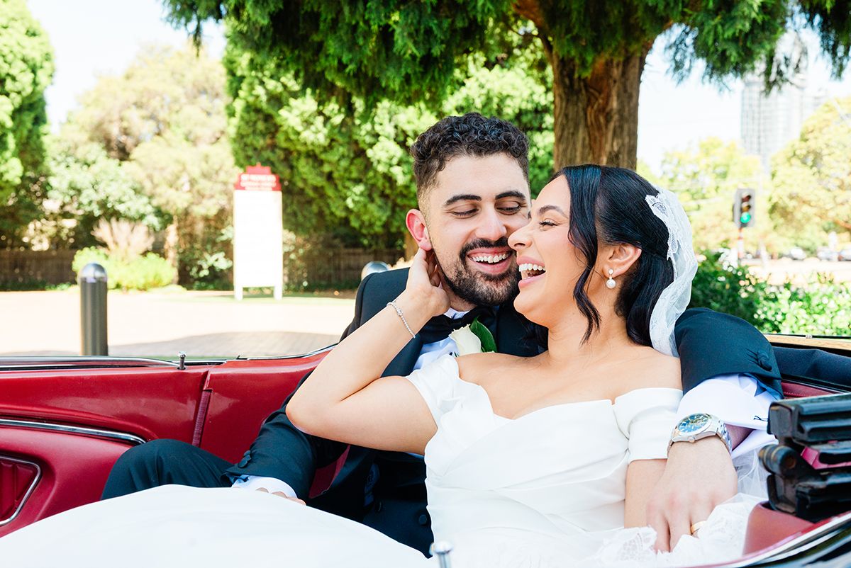 Bride and groom smiling and embracing in a red convertible car on their wedding day.