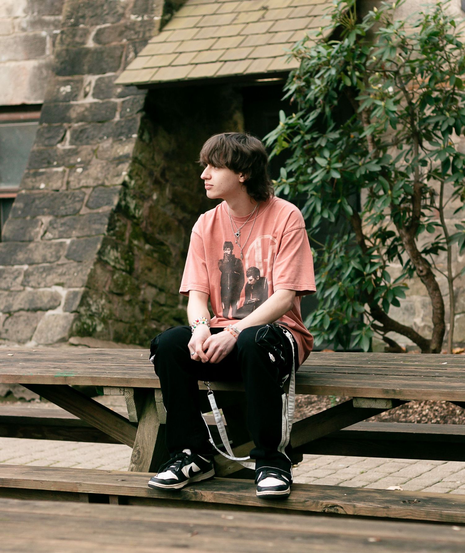Young person in pink graphic tee and black pants sits on wooden bench outdoors near stone building.