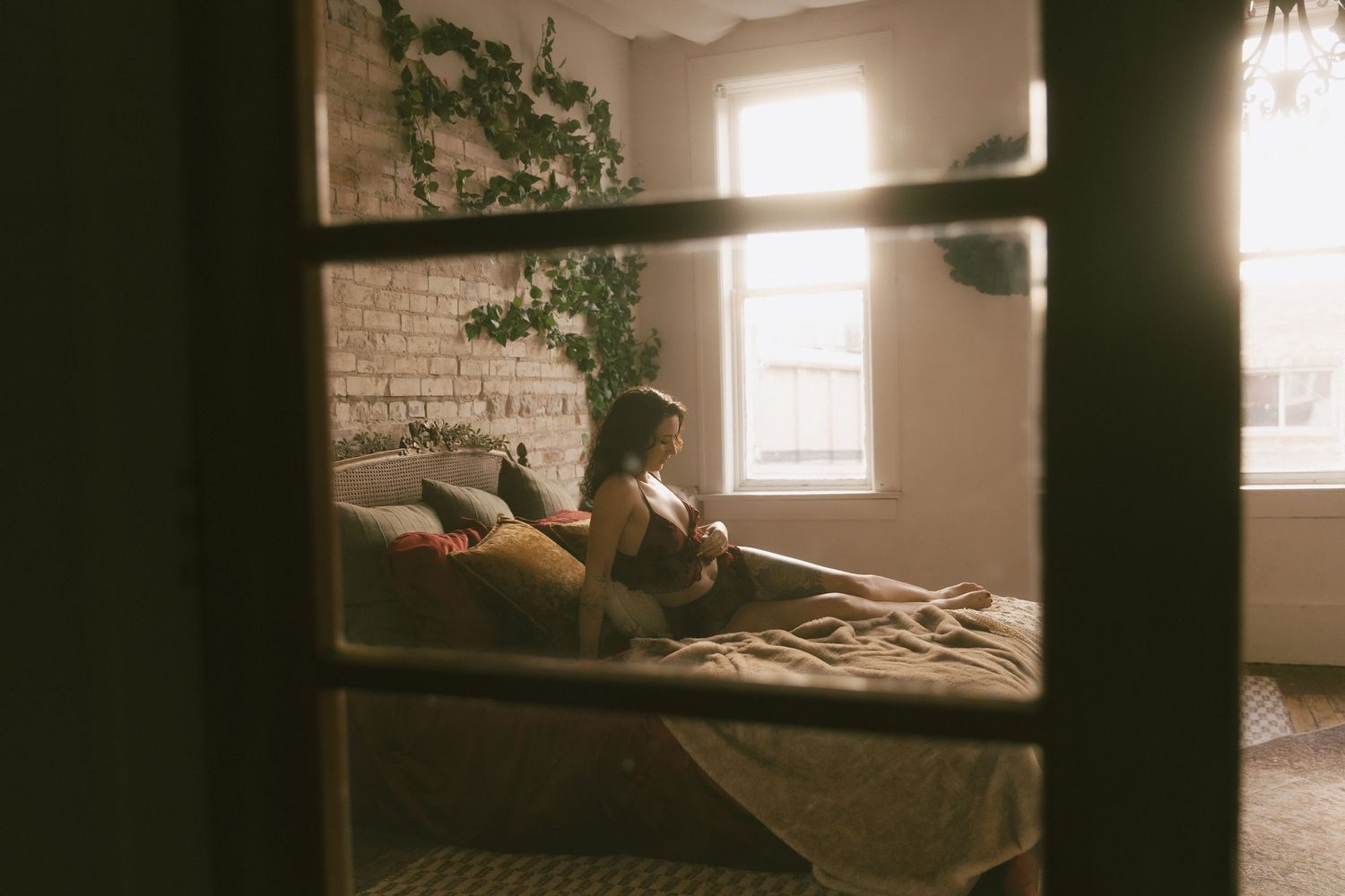 Woman relaxing on a bed with a cat, sunlit bohemian bedroom with brick wall and hanging plants.