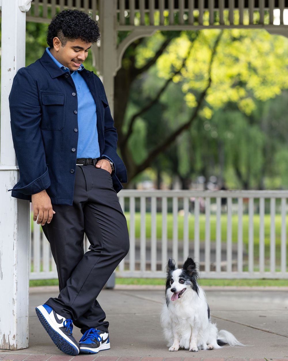 A young man in a navy jacket and blue sneakers leans against a white post, looking down at a black and white Border Collie.