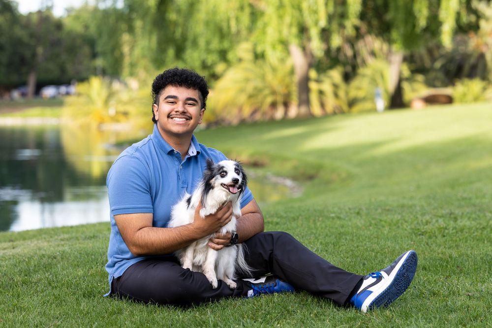Young man smiling outdoors on green grass, holding a small black and white dog near a pond.