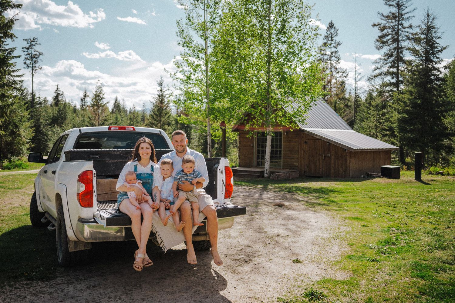 Family of four sitting on truck tailgate near rustic log cabin surrounded by pine trees on sunny day.