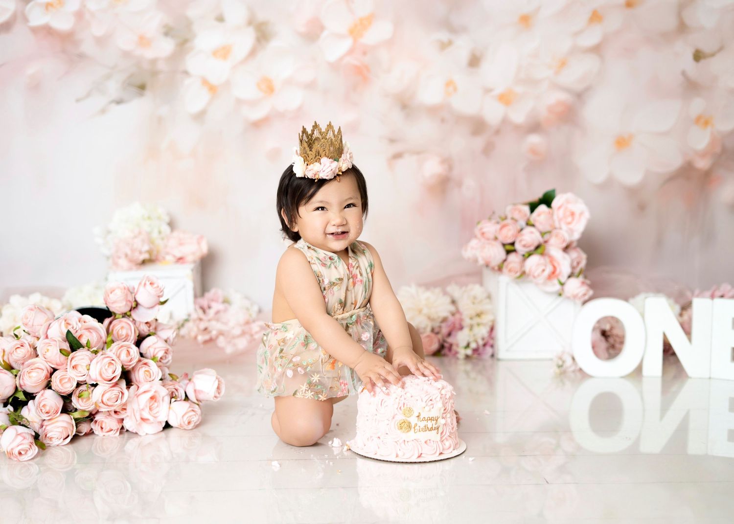 Baby girl first birthday cake smash with gold crown, pink floral setup, roses and “ONE” decor in Covina studio