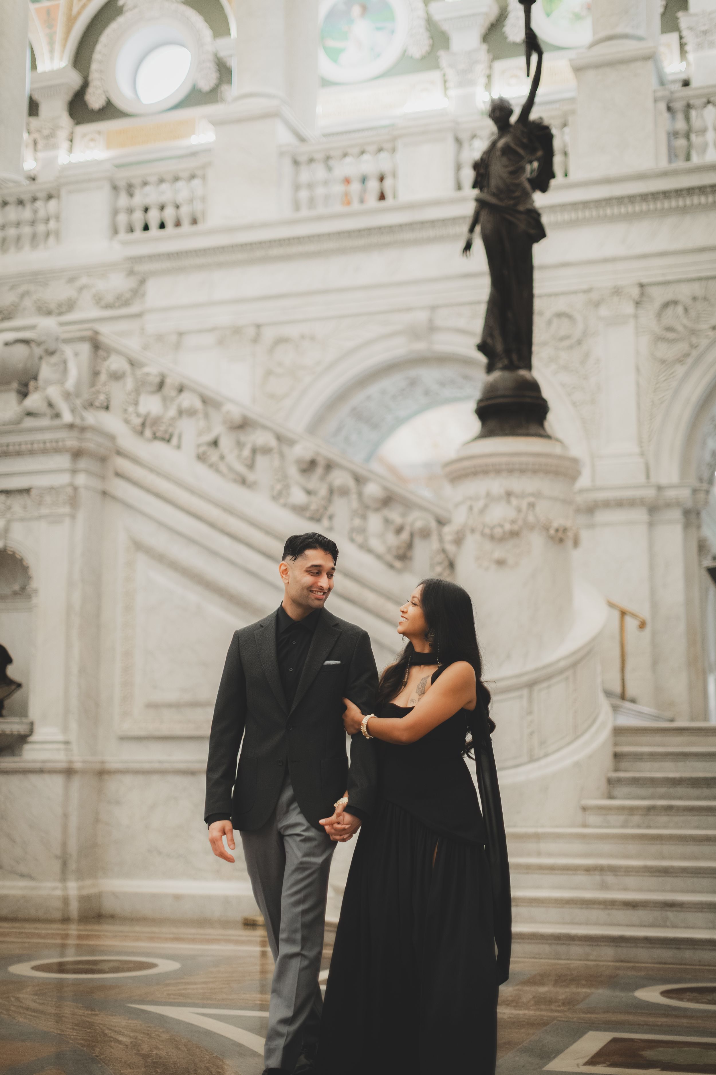 Couple in elegant black attire walking hand-in-hand inside a grand marble building with ornate staircases.