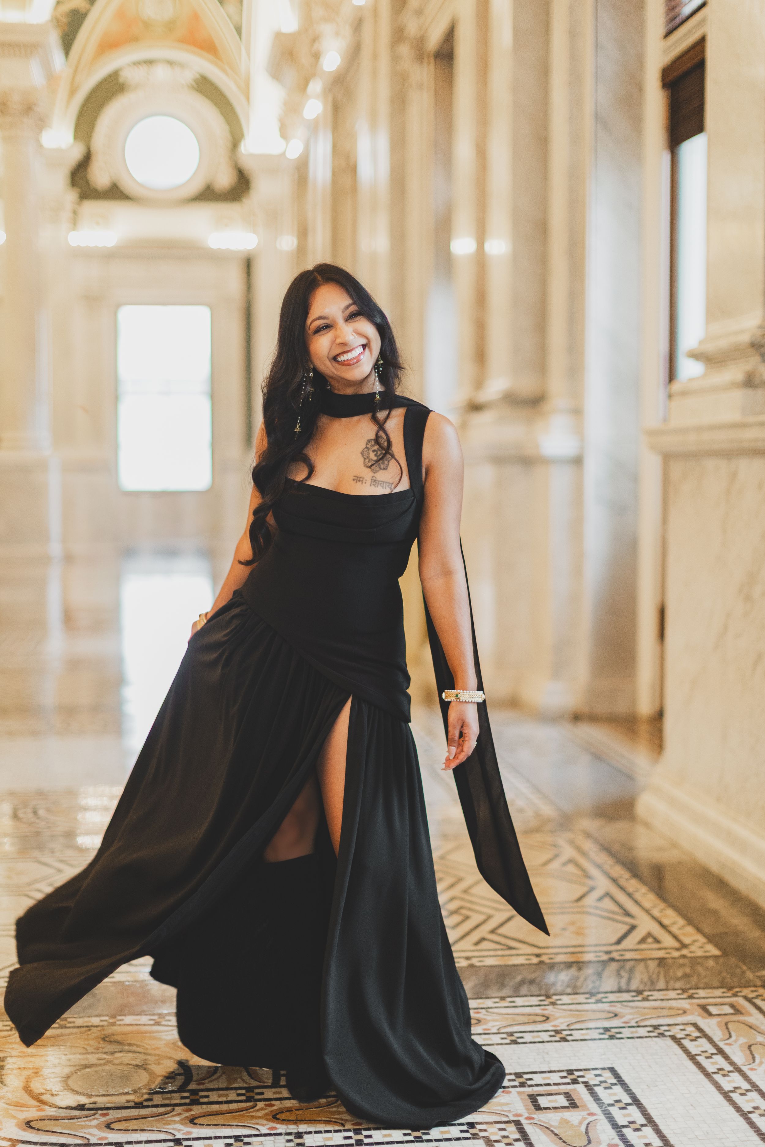 Woman in elegant black gown with slit poses gracefully in ornate marble hallway, smiling confidently captured by stefan a photography