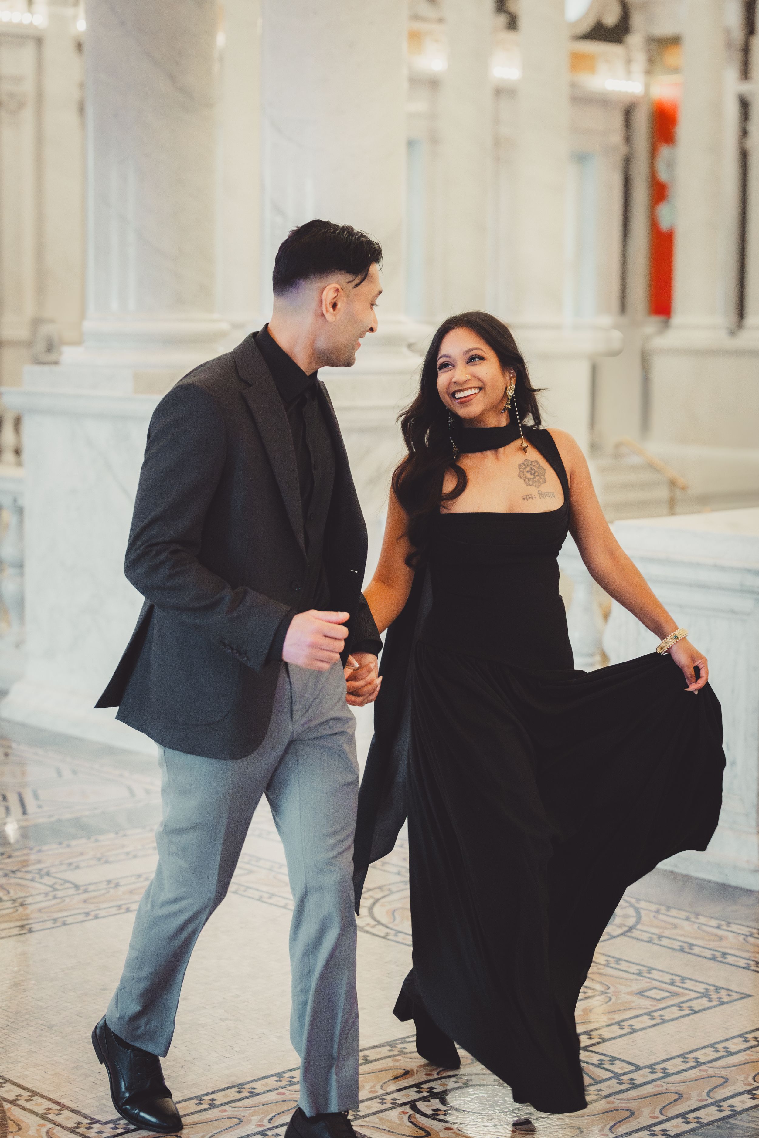 Elegant couple in black formal attire walking hand in hand through a grand marble hall, smiling at each other.