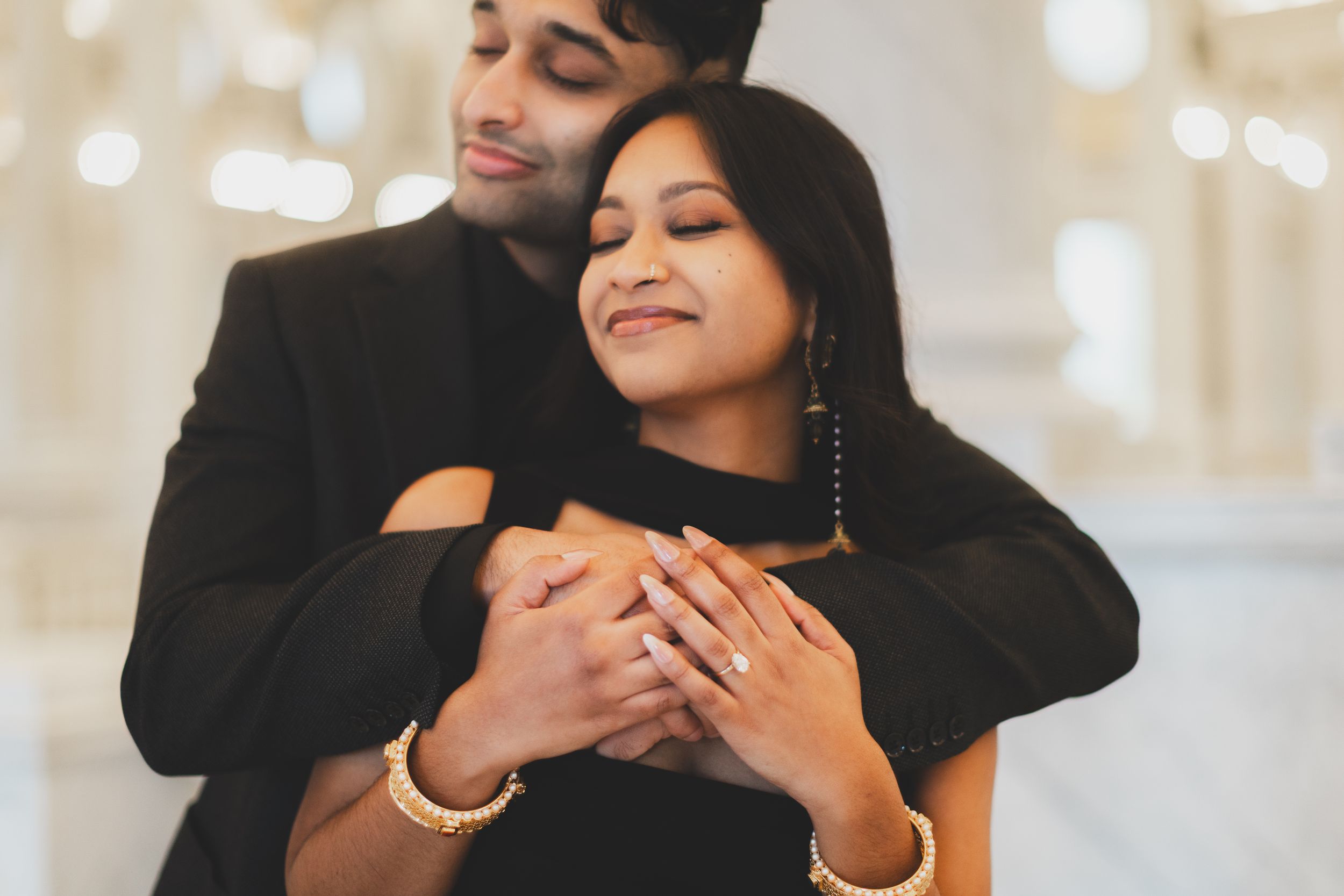 Couple in black outfits sharing a romantic embrace, woman wearing gold bangles and engagement ring. Captured by a dc engagement photographer