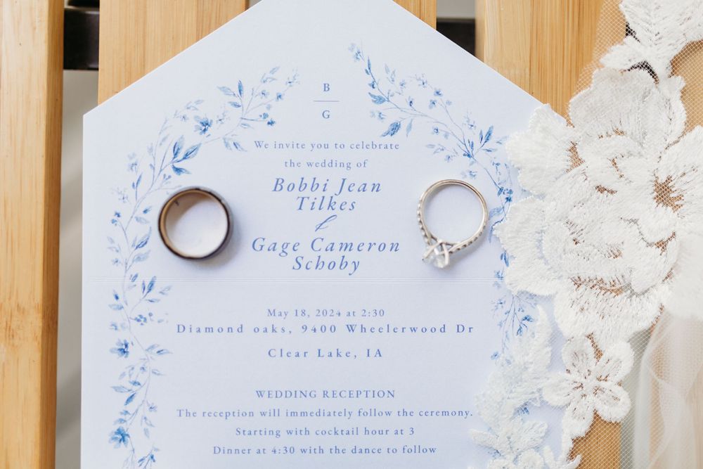 Wedding invitation for Bobbi Jean Tilkes and Gage Cameron Scoby displayed with gold wedding bands and white floral details.