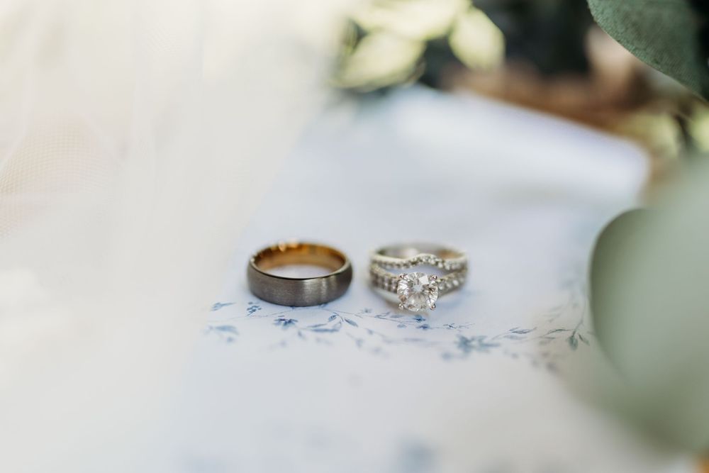 Two wedding rings, a gold tungsten band and a diamond engagement ring, rest on a soft blue floral surface.