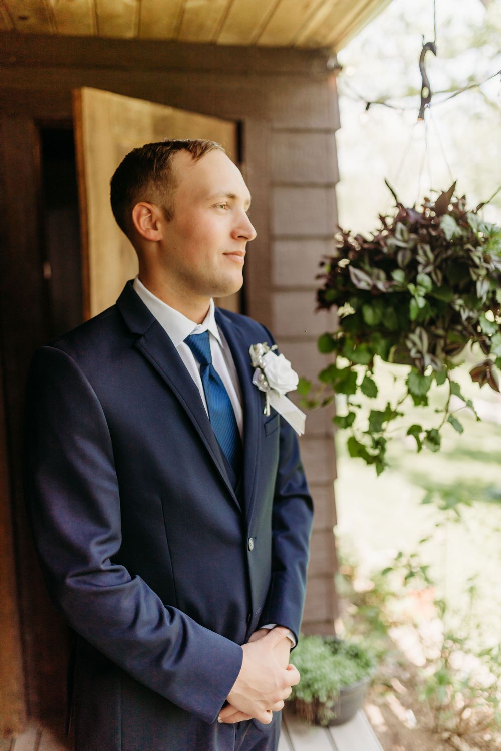 Groom in navy blue suit with white boutonniere stands on wooden porch, gazing thoughtfully to the side.