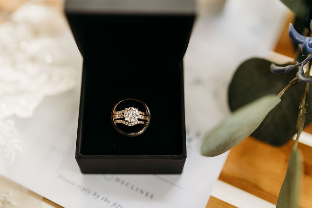 Gold diamond engagement ring displayed in a black velvet jewelry box, surrounded by elegant wedding stationery and greenery.