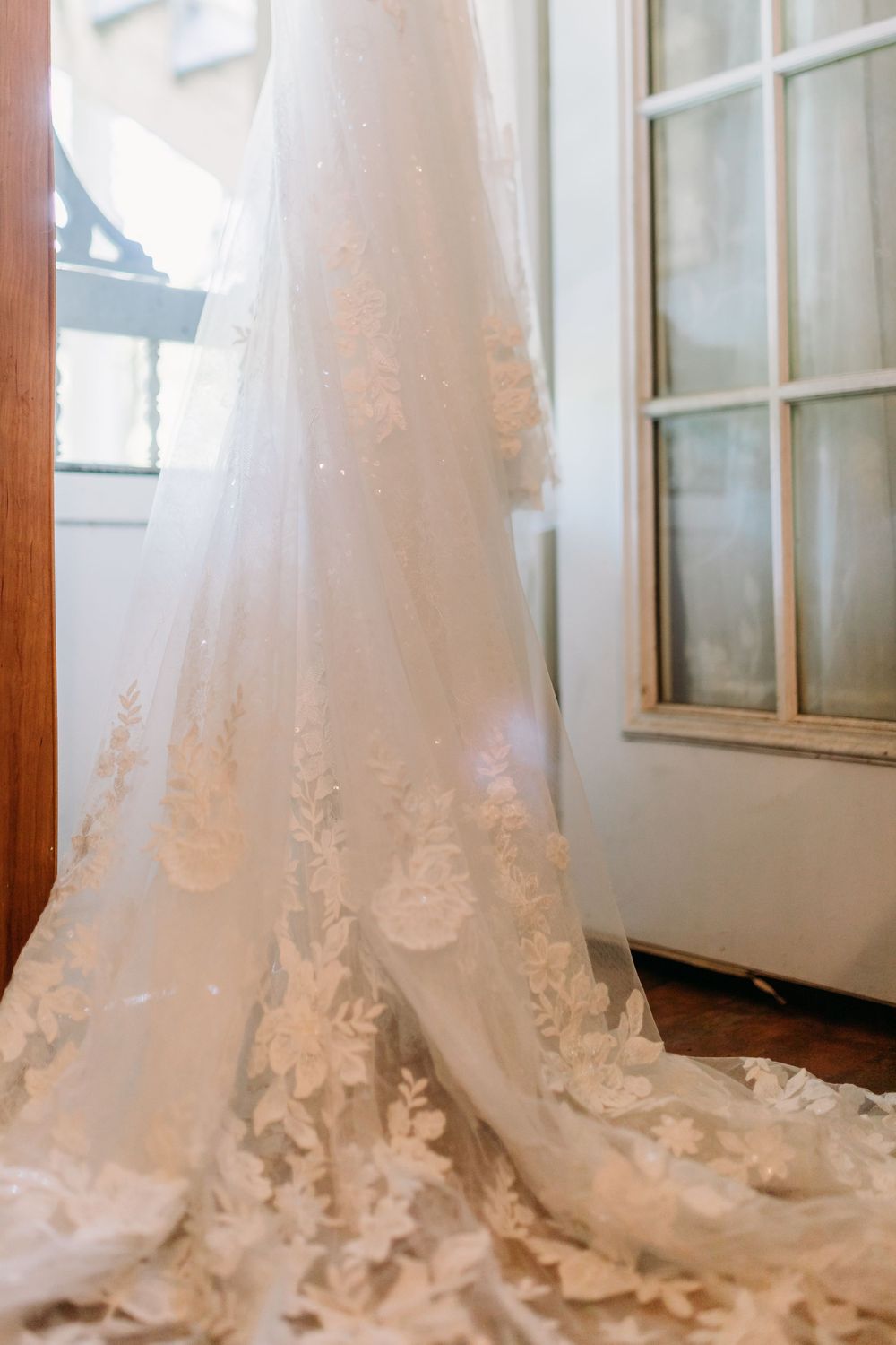 Elegant white lace wedding gown with floral appliques and a flowing train hanging near a vintage window.