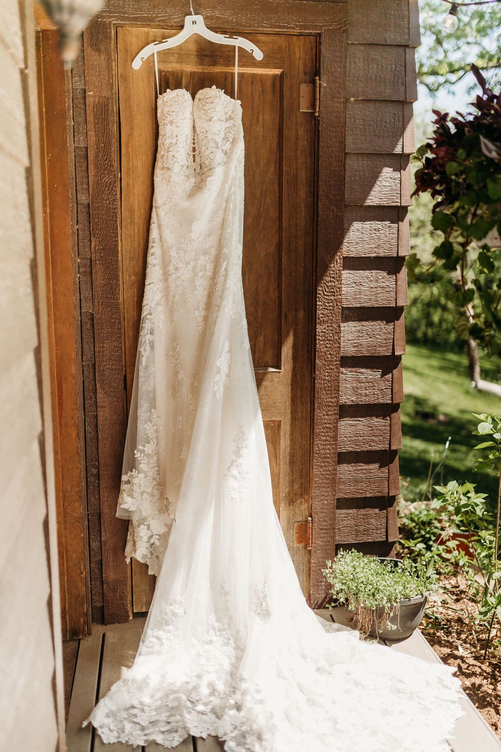 Elegant white lace wedding dress hanging on a wooden door in natural sunlight at an outdoor venue.