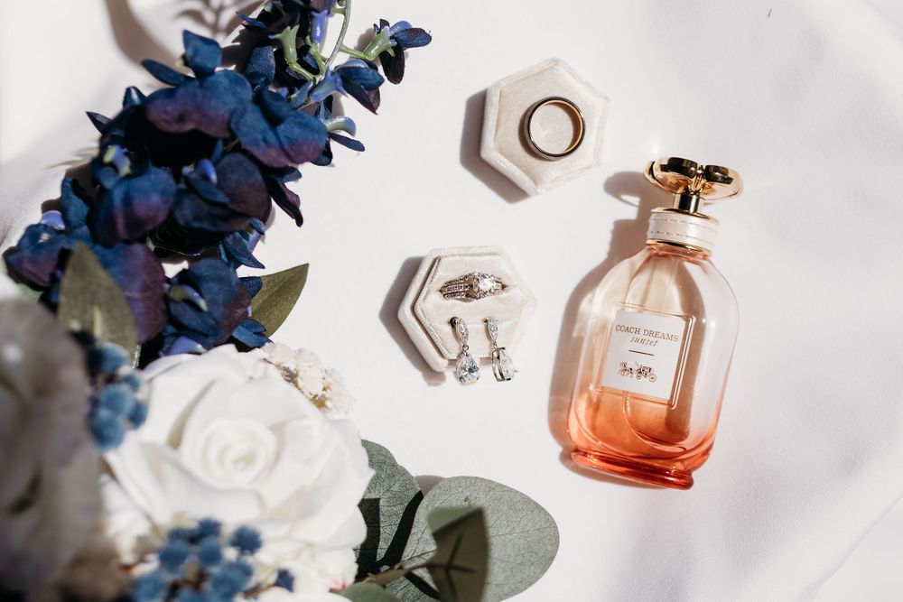 Elegant wedding flat lay with peach perfume bottle, gold rings, earrings, and blue and white floral bouquet on white fabric.