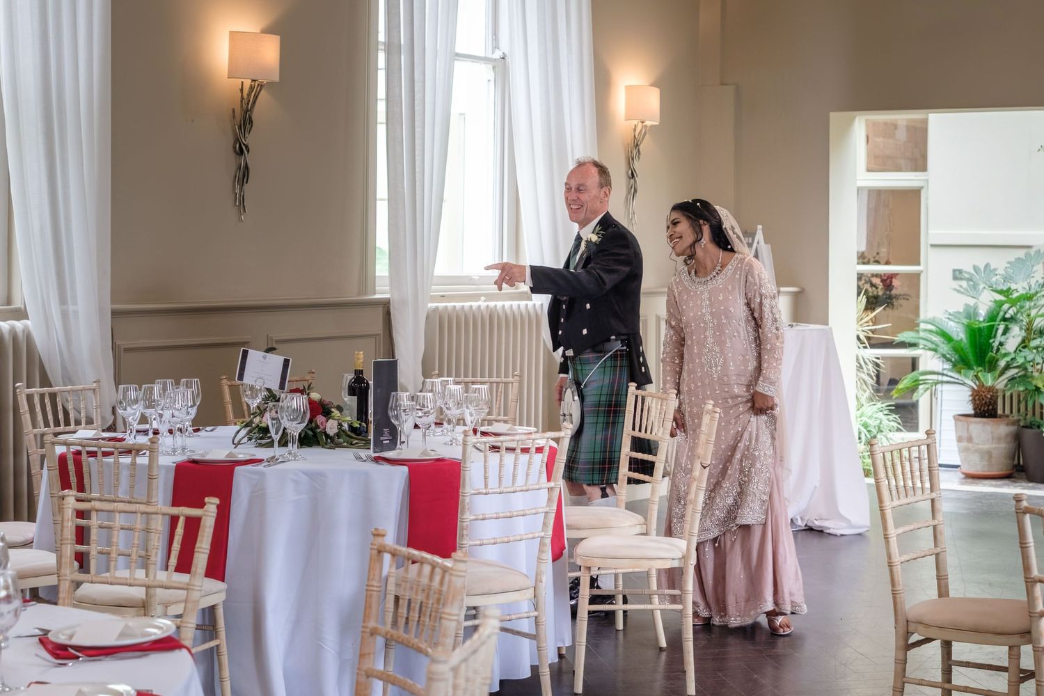 Elegant wedding reception venue with round tables, red floral centerpieces, and a couple conversing near large windows.