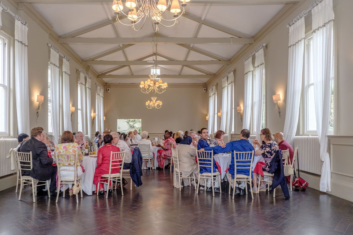 Elegant wedding reception in a grand hall with chandeliers, guests seated at round tables with colorful chairs.
