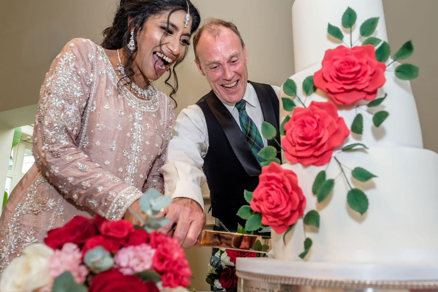 Bride and groom laughing while cutting a white wedding cake decorated with red roses.