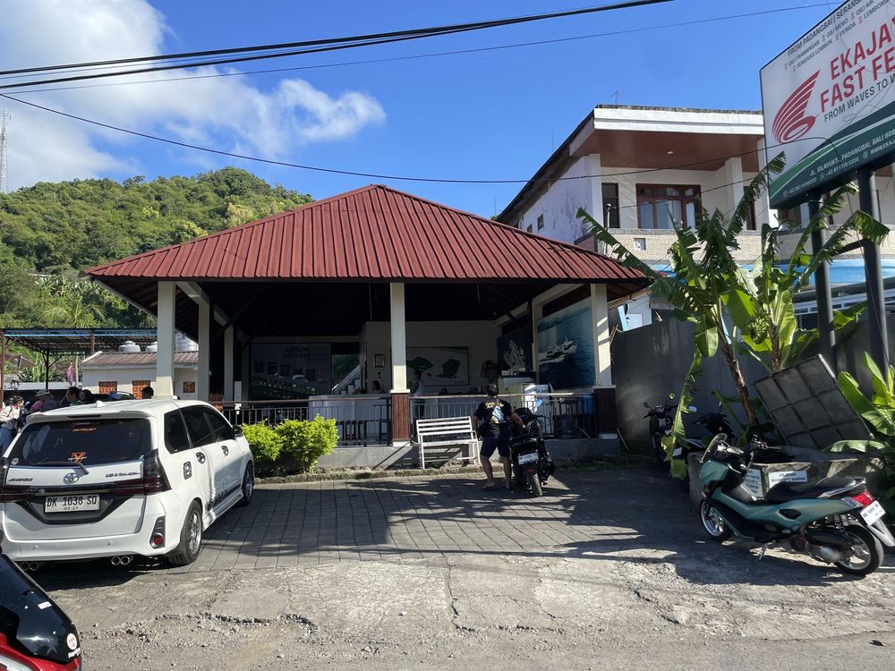 Small Indonesian roadside restaurant with red roof, parked cars, motorcycles, and lush green hills in the background.