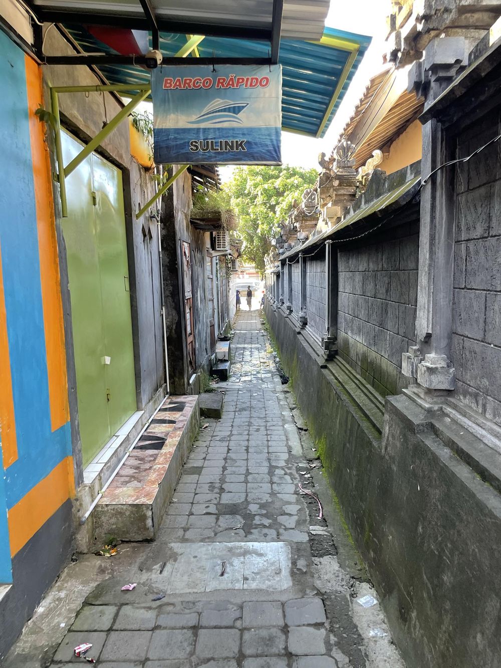 Narrow cobblestone alley in Bali Indonesia with traditional stone walls and a Marco Marino Sulink sign overhead.