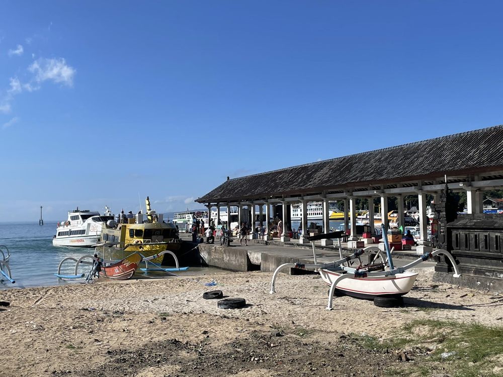 Traditional outrigger boats on a rocky beach beside a waterfront building with ferries docked under a clear blue sky.