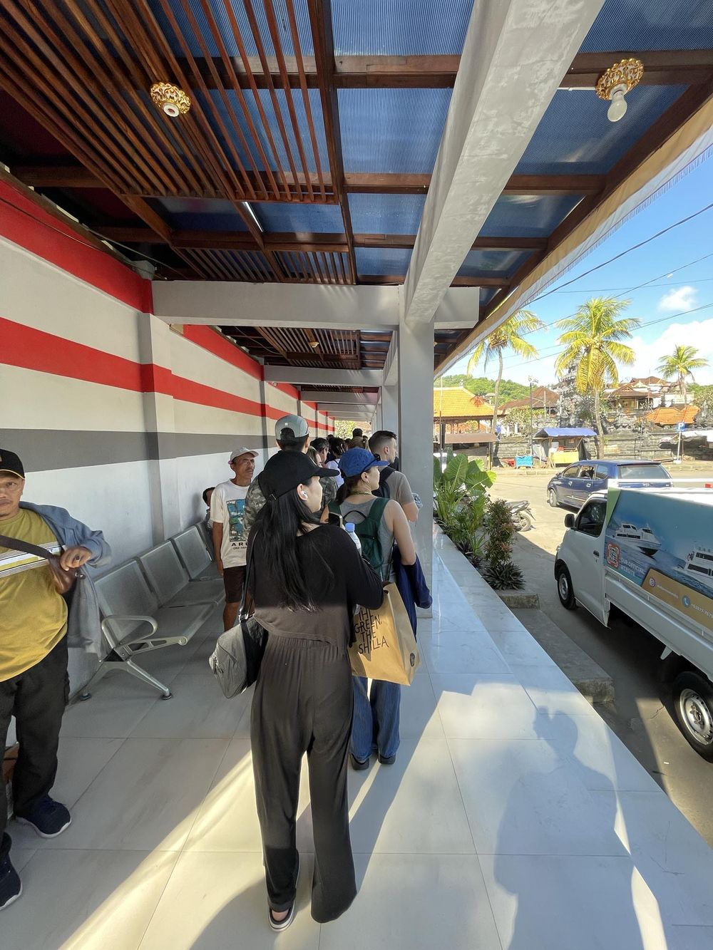 People queue outside a modern covered walkway with wooden ceiling and red stripe wall in a tropical setting.