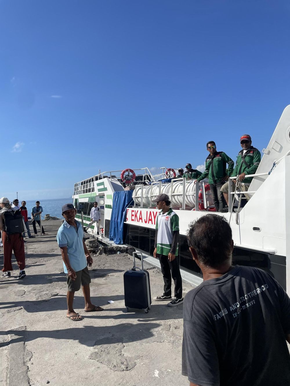Passengers board the Eka Jaya ferry boat at a sunny tropical harbor pier with luggage.