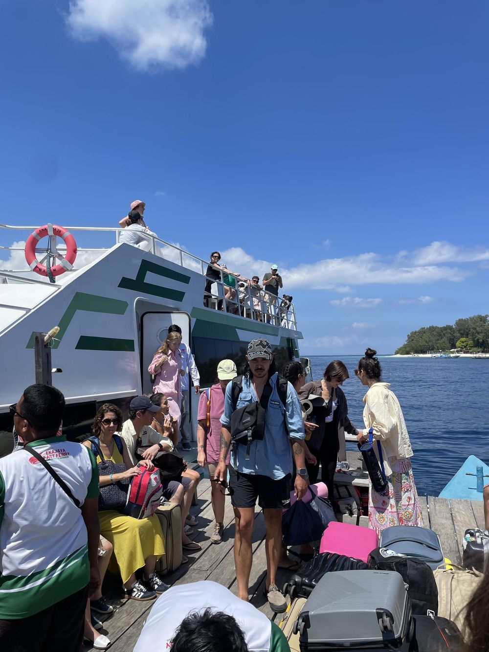 Tourists disembarking a ferry boat onto a wooden dock with tropical blue ocean and island in the background.