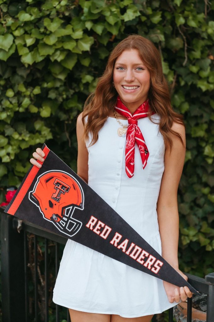 Smiling young woman in white dress holds Texas Tech Red Raiders pennant with red bandana scarf outdoors.