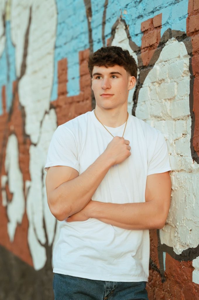 Young man in white t-shirt and jeans poses confidently against a colorful graffiti brick wall.