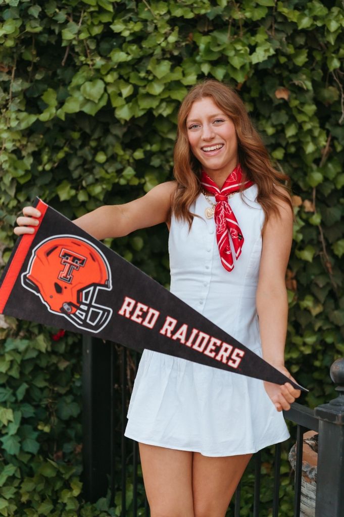Smiling young woman holding a Texas Tech Red Raiders pennant flag, wearing a white dress and red bandana outdoors.