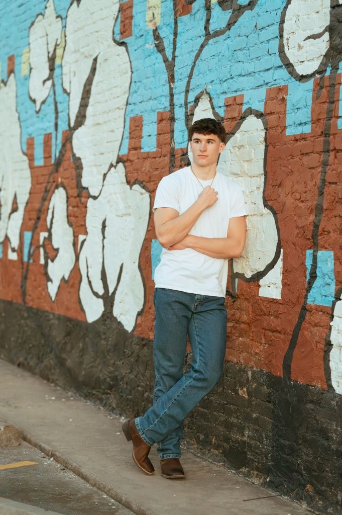 Young man in white t-shirt and jeans leaning against a colorful graffiti brick wall outdoors.