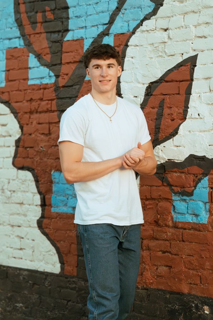 Young man in white t-shirt and jeans smiling against colorful graffiti brick wall backdrop.