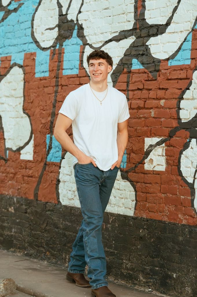Smiling young man in white t-shirt and jeans leaning against colorful graffiti brick wall.