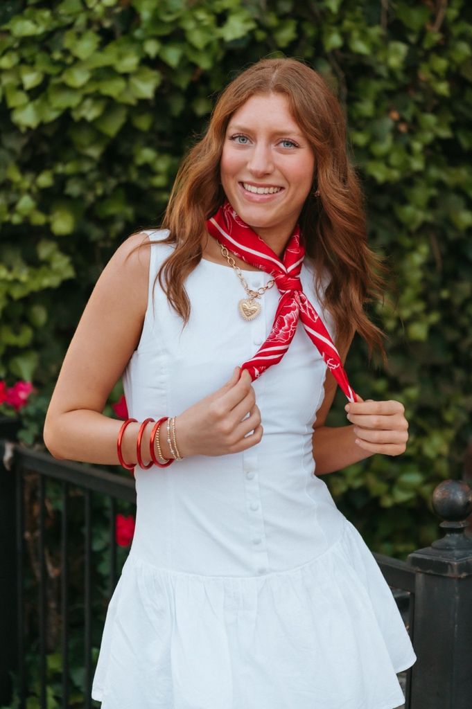 Close-up of smiling woman in white dress with red bandana scarf, gold heart necklace, and red bangles outdoors.