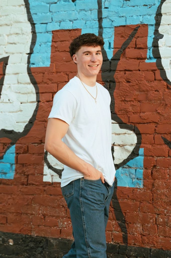 Young man in white t-shirt and jeans smiling sideways against vibrant graffiti brick wall.