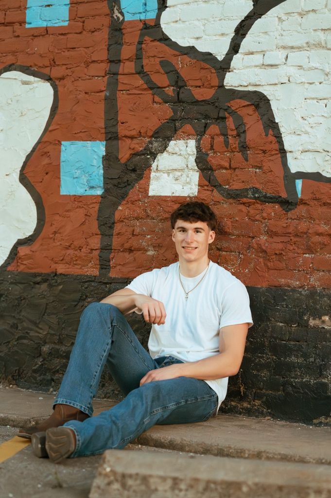 Young man in white t-shirt and jeans sitting relaxed against colorful graffiti brick wall outdoors.