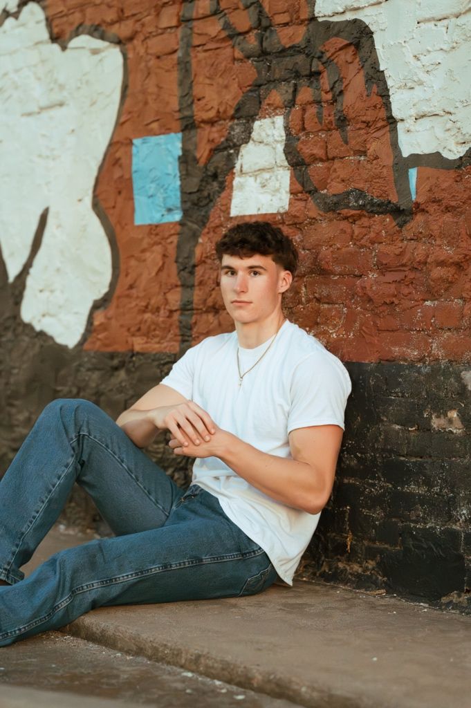Young man in white t-shirt and jeans sitting against a graffiti brick wall in a relaxed pose.
