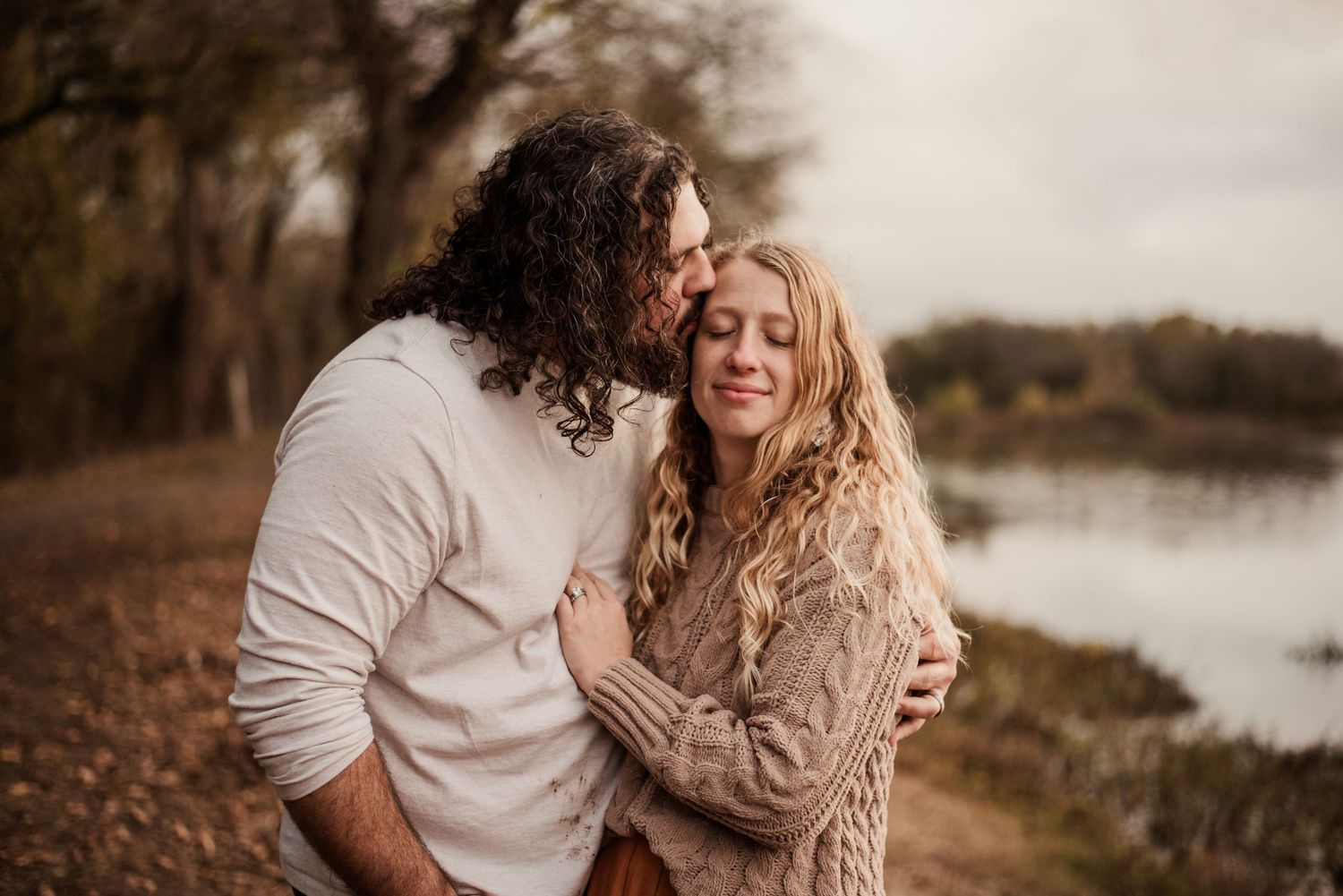 Couple embracing outdoors near a lake in autumn, man with curly hair kissing woman's cheek as she smiles with eyes closed.