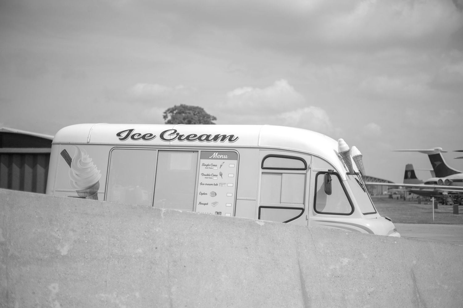 Vintage black and white photo of a classic ice cream van parked near an airfield on a cloudy day.