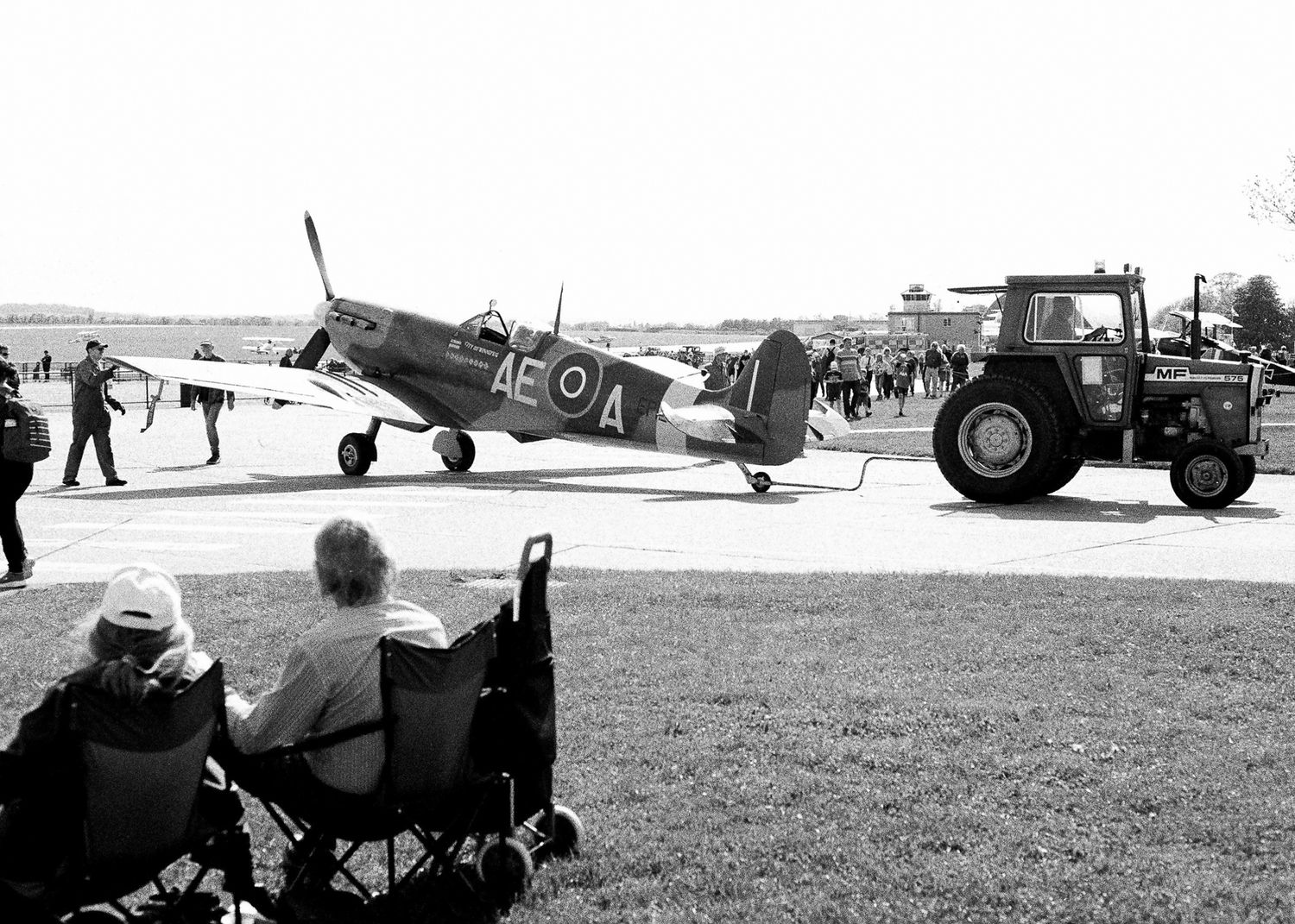 Black and white photo of a vintage WWII Spitfire aircraft being towed by a tractor at an airshow with spectators watching.