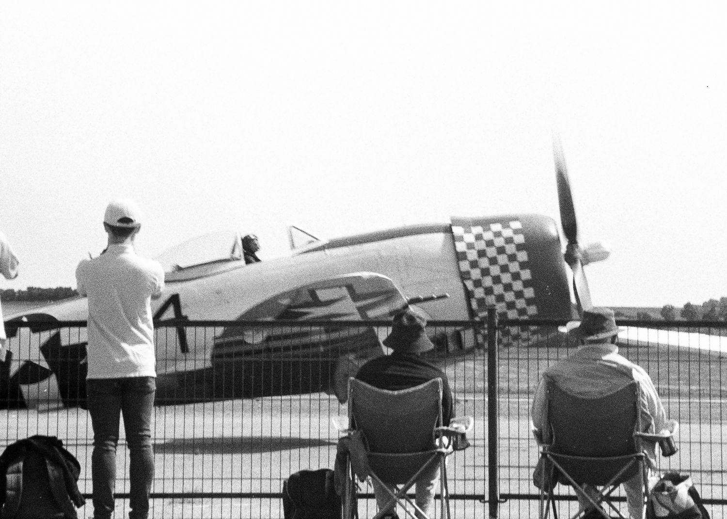 Black and white photo of spectators watching a vintage P-47 Thunderbolt warplane at an airshow behind a fence.