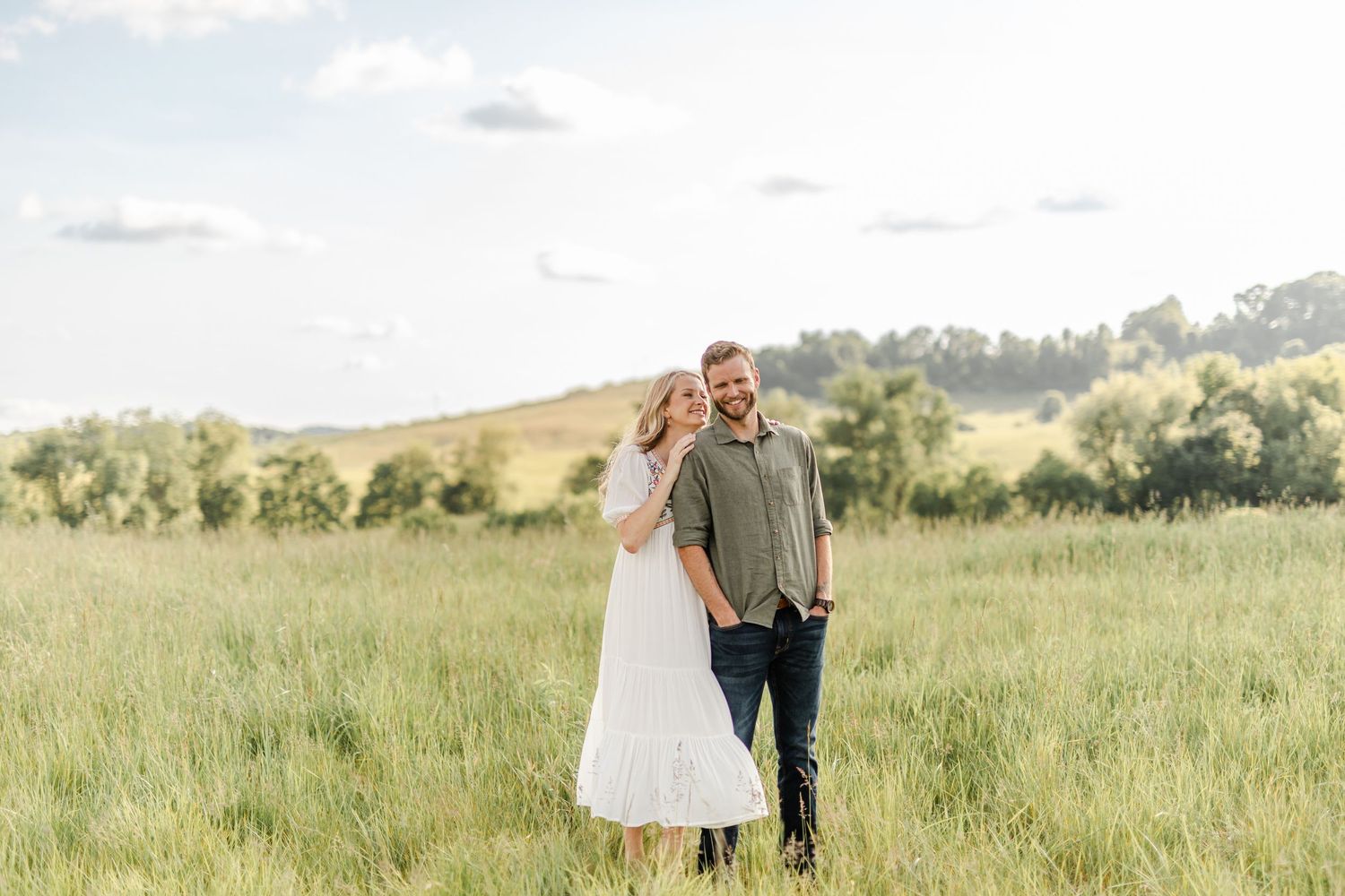 Couple embracing in lush green meadow under bright sky during outdoor photo session.