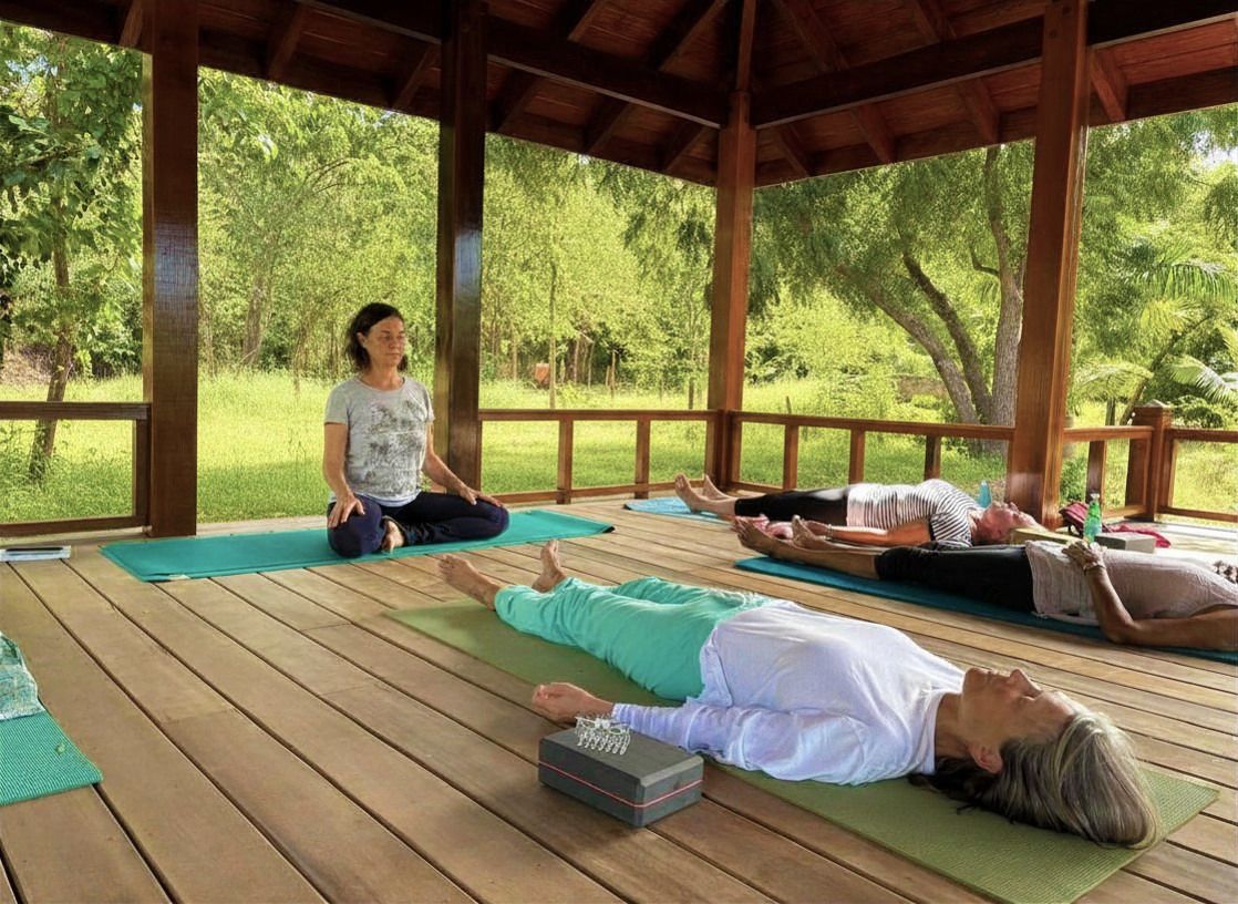 Yoga instructor leads a relaxation class on a wooden pavilion with green mats surrounded by lush trees.